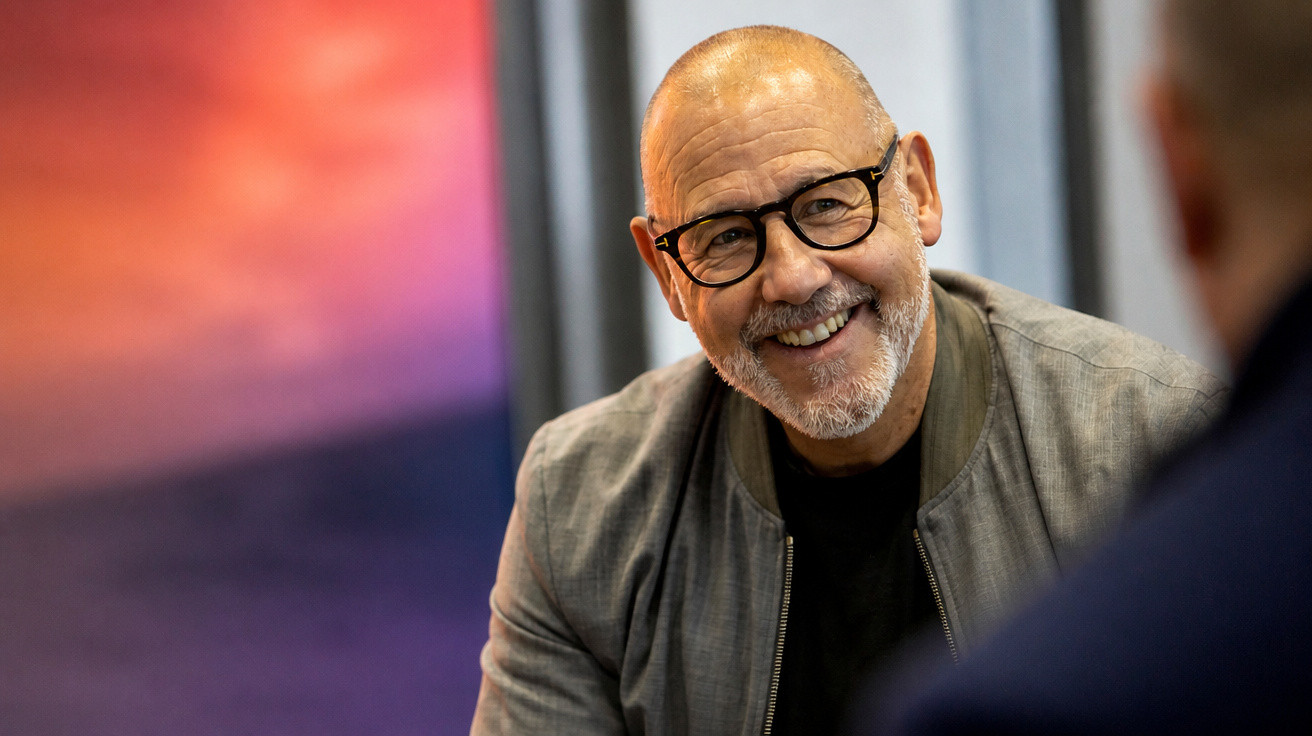 Smiling bald man with grey beard and glasses wearing a grey jacket, engaged in conversation indoors.
