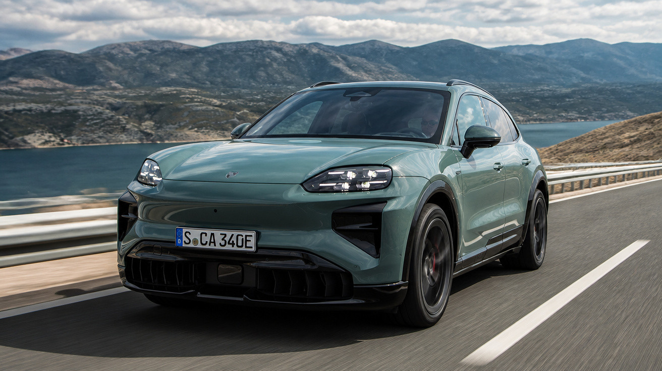Green electric SUV driving on coastal road with mountains and water in the background under cloudy sky.